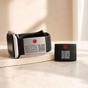 May include: A black smartwatch band featuring a silver-toned medical alert tag with a red Star of Life symbol, a QR code, and the text: Lovis A. Paterson, Miami, FL - USA, Contact phone #1, Contact phone #2. A matching black box is placed nearby.