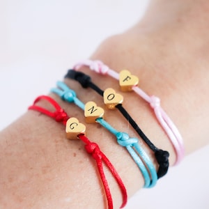 May include: Five colorful friendship bracelets with gold heart charms, each charm has a letter engraved on it, spelling out the word 'FONG'.