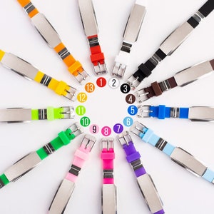 K&ouml;nnte beinhalten: Ein Set mit 13 bunten Silikonarmb&auml;ndern mit silbernen Metallplatten. Jedes Armband hat eine Nummer von 1 bis 13.