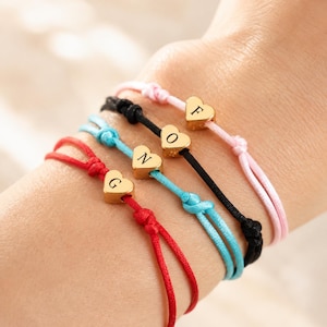 Puede incluir: Cuatro pulseras de cordón en rojo, azul claro, negro y rosa claro. Cada pulsera cuenta con un dije dorado en forma de corazón. Los dijes están grabados con las letras G, N, O y F, centrados en el cordón mediante nudos decorativos.