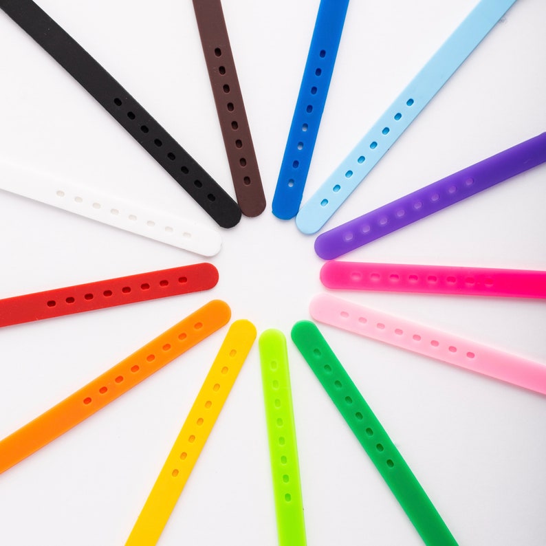 K&ouml;nnte beinhalten: Eine Sammlung von 12 bunten Gummib&auml;ndern, die in einem Kreis auf wei&szlig;em Hintergrund angeordnet sind. Die B&auml;nder sind in verschiedenen Farben, darunter Rot, Orange, Gelb, Gr&uuml;n, Blau, Lila, Rosa und Schwarz.