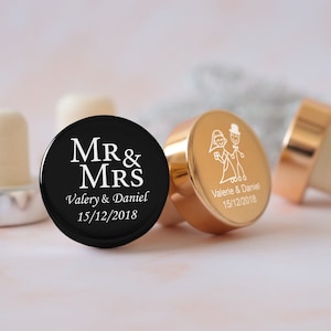 May include: Two personalized bottle stoppers, one black with white text "MR & MRS Valery & Daniel 15/12/2018" and one gold with a white illustration of a bride and groom and the text "Valerie & Daniel 15/12/2018".