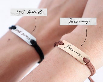 May include: Two bracelets with personalized messages. One bracelet has a black cord and a silver plate with the handwritten message "LOVE ALWAYS". The other bracelet has a brown cord and a gold plate with the handwritten message "Grammy".