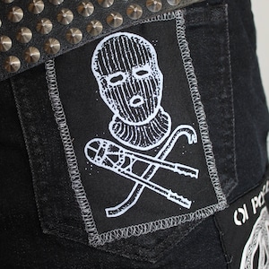 Puede incluir: Vaqueros negros de denim con un parche negro y blanco que representa una figura enmascarada y herramientas cruzadas. Los vaqueros tienen un cinturón tachonado.