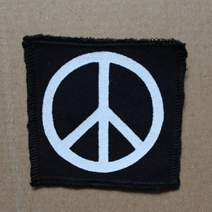 Könnte beinhalten: Ein schwarzer Stoff-Patch mit einem weißen Peace-Zeichen in der Mitte.