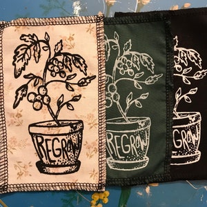 Può includere: Tre toppe di tessuto con un'illustrazione in bianco e nero di una pianta in vaso con la parola "REGROW" stampata sul vaso. Le toppe sono di diversi colori: beige, verde e nero.