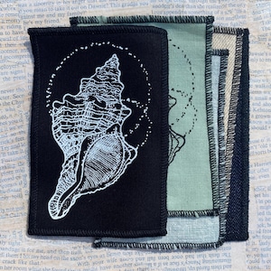 May include: Four fabric patches featuring a white seashell design on black, sage green, and navy blue. The patches have stitched edges and are on a backdrop of old book pages.