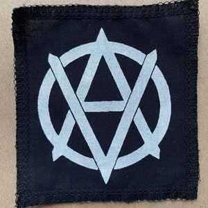 Op de afbeelding: Een zwarte stoffen patch met een witte cirkel en een witte driehoek in de cirkel. De driehoek heeft een punt bovenaan en twee punten onderaan. De cirkel is rond de driehoek.