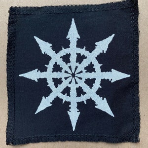 Op de afbeelding: Een zwarte stoffen patch met een wit achtpuntig sterontwerp. De ster heeft een cirkel in het midden met kleinere cirkels eromheen.