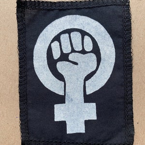 Può includere: Toppa in tessuto nero con un simbolo femminista bianco, un pugno chiuso all'interno di un cerchio sopra una croce. La toppa ha un bordo nero cucito.