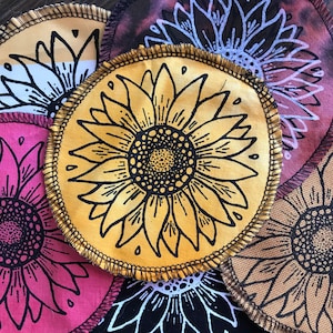 Könnte beinhalten: Ein Set mit sechs Stoffuntersetzern mit einem Sonnenblumenmuster. Die Untersetzer sind rund und haben ein schwarz-gelbes Farbschema. Die Sonnenblumen sind schwarz umrandet und haben einen gelben Mittelpunkt.