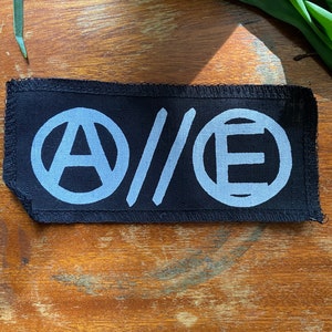 Peut inclure: Patch en tissu noir avec un symbole d'anarchie brodé en blanc, une double barre oblique et un cercle avec la lettre E.