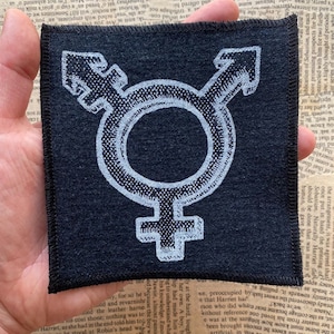 May include: A white embroidered transgender symbol on a black fabric patch.