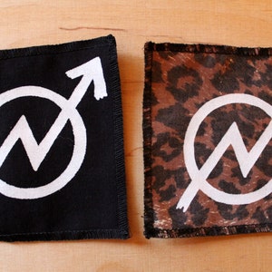 Könnte beinhalten: Zwei schwarze Stoffpatches mit weißen Nähten an den Rändern. Jeder Patch zeigt einen weißen Kreis mit einem weißen Blitz darin und einem weißen Pfeil, der nach rechts zeigt. Ein Patch hat einen schwarzen Hintergrund und der andere einen braunen Leopardenmuster-Hintergrund.
