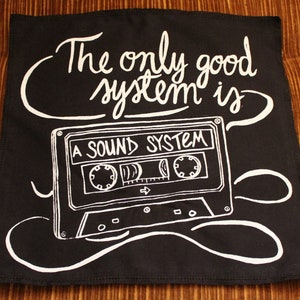 Könnte beinhalten: Schwarzer quadratischer Stoff mit weißem Text und einer Kassettenband-Illustration. Der Text lautet "The only good system is A SOUND SYSTEM". Das Kassettenband-Design enthält Etiketten und Knöpfe.