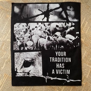 Op de afbeelding: Zwart-witte patch met een grafisch ontwerp van een vogel, een prikkeldraadhek en een groep dieren. De tekst "YOUR TRADITION HAS A VICTIM" is op de patch gedrukt.