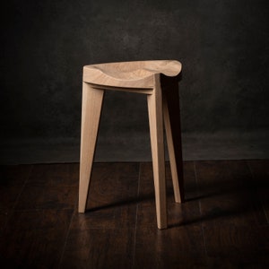 Könnte beinhalten: Ein Hocker aus Holz mit einer geschwungenen Sitzfläche und vier Beinen. Der Hocker ist aus hellbraunem Holz gefertigt und hat eine natürliche Oberfläche.