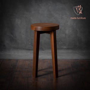 K&ouml;nnte beinhalten: Ein massiver Holzhocker mit runder Oberseite und vier Beinen. Der Hocker ist aus dunkelbraunem Holz gefertigt und hat ein schlichtes, modernes Design.