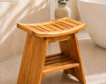 May include: A light brown wooden shower stool with a curved, slatted seat and a lower storage shelf. The frame features angled legs and the text meda furniture is engraved on the front support rail, showcasing a natural wood grain finish.