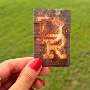 May include: A rectangular card featuring a dark brown wood grain background with a large, stylized letter R outlined in barbed wire. The letter is filled with a vibrant orange and yellow flame pattern, complete with glowing embers and sparks.