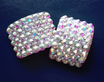 May include: Two iridescent, crystal-covered accessories, possibly belt buckles, are displayed against a dark blue background. The crystals reflect a spectrum of colors, including pink, purple, and silver, creating a dazzling effect. The accessories have a rectangular shape.
