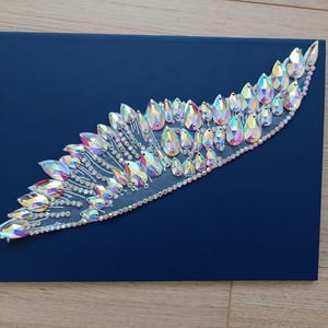 May include: A clear iridescent crystal embellishment in the shape of a wing, with a teardrop design. The embellishment is on a dark blue background.