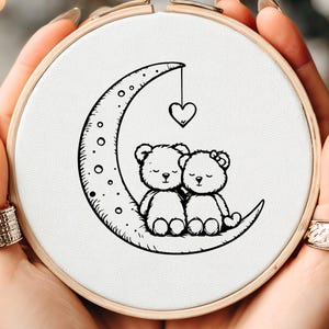 May include: An embroidery hoop with a black and white design of two teddy bears on a crescent moon, with a hanging heart. The design is on white fabric, held in a wooden hoop.