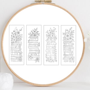 Pode incluir: Quatro marcadores de livros a preto e branco com pilhas de livros adornadas com arranjos florais. Os marcadores de livros são apresentados dentro de um bastidor de bordado de madeira. A imagem é um close-up, destacando os detalhes das ilustrações.