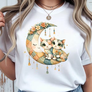 Puede incluir: Camiseta blanca con una ilustración caprichosa de dos gatos abrazados en una luna creciente. La luna está decorada con un diseño de patchwork y detalles florales. Los gatos tienen ojos verdes y están adornados con flores.