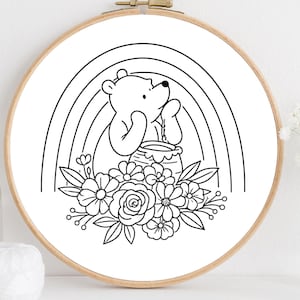 May include: A black and white line drawing of a bear holding a honey pot, set against a rainbow and floral arrangement, framed in a wooden embroidery hoop. The design is suitable for embroidery or coloring.