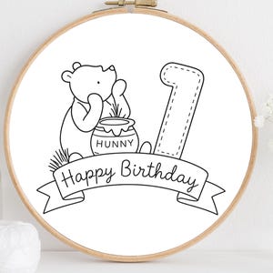 May include: A black and white illustration of Winnie the Pooh with a honey pot and the number one inside a wooden embroidery hoop. The words "Happy Birthday" are written on a banner below the image.