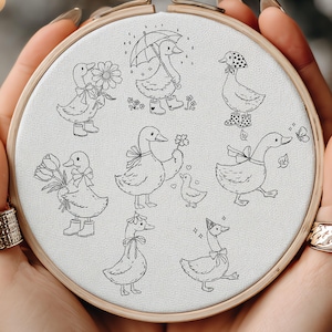 May include: An embroidery hoop with a collection of charming duck illustrations in black line art. The ducks are depicted in different outfits and activities, such as holding flowers or wearing boots. The artwork is set on a neutral fabric.