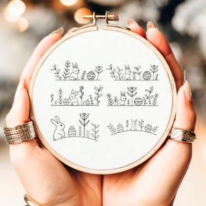 May include: A wooden embroidery hoop holds white fabric featuring six black line drawings of Easter bunnies, eggs, and floral elements. The designs are arranged in horizontal rows, creating a decorative border. The hoop is held by hands.