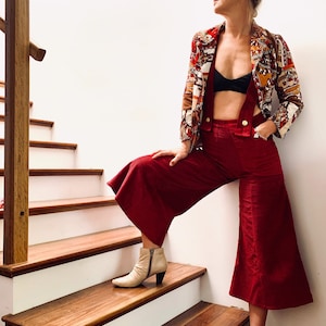 May include: A woman wearing a red corduroy jumpsuit with a wide-leg silhouette. She is also wearing a patterned blazer and white heeled boots. The woman is standing on a wooden staircase with white risers.