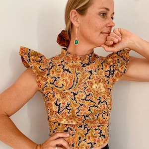 May include: A woman wearing a black and gold paisley print top with ruffled sleeves. The top has a high neck and a fitted silhouette.