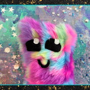 May include: A colorful, furry creature with a smiling face and large black eyes. The plush toy features a mix of pink, purple, blue, and green hues. The background is a dreamy mix of colors with stars and glitter.