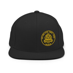 Può includere: Cappellino nero a visiera piatta con un emblema giallo. L'emblema circolare presenta un occhio in un triangolo, circondato dal testo "LUCKY PRICK TATTOO - PHOENIX - ARIZONA" e un numero di telefono.