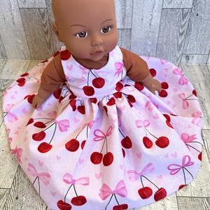 May include: A doll wearing a pink dress with a cherry and pink bow print. The doll has brown skin and brown eyes.