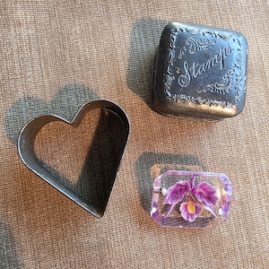 May include: An assortment of vintage items on a brown surface. Includes a heart-shaped cookie cutter, a small metal stamp holder with the word "Stamps", and a clear resin paperweight with a purple flower.