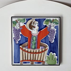 May include: A square ceramic tile featuring a hand-painted illustration of a figure in a red tunic standing inside a wooden barrel. The figure holds a cluster of purple grapes in each hand against a deep blue background with green foliage.