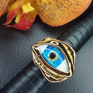 May include: A gold-colored ring with an eye-shaped design. The eye features a blue iris with yellow and black details, set against a white background. The ring is on a black, textured surface.