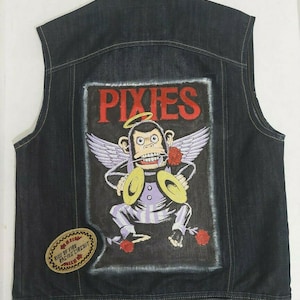 May include: A dark blue denim vest with a large embroidered patch on the back. The patch features a cartoon monkey with angel wings playing cymbals. The text "PIXIES" is above the monkey in red lettering. A smaller patch with the text "RING OF FIRE RACECAR CIRCUIT" is on the left side of the vest.