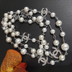 May include: A long necklace featuring a silver-toned chain interspersed with white faux pearls of varying sizes. The design includes multiple silver-toned CC logos encrusted with small clear crystals, spaced evenly throughout the length of the chain.