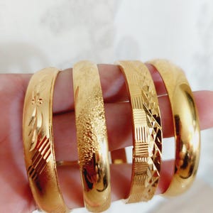 May include: Four gold bangles with different engraved patterns. The bangles are stacked on top of each other.