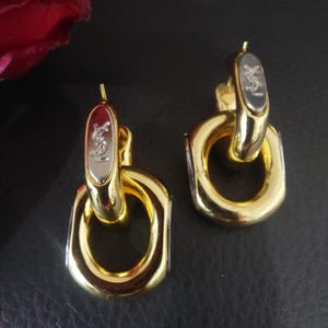 May include: A pair of gold-tone metal earrings featuring a dangling oval hoop design. Each earring has a silver-tone top accent engraved with the YSL logo. The earrings have a polished, reflective finish and a post-back closure mechanism.