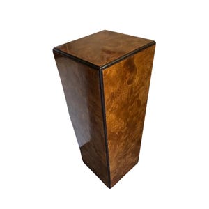 May include: A tall, rectangular pedestal with a glossy, burl wood finish. The top and edges are outlined with a dark trim. The pedestal has a trapezoidal shape, wider at the top and tapering towards the base. The wood grain is visible.