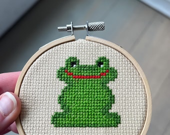 Könnte beinhalten: Ein grünes Frosch-Kreuzstich-Design in einem hölzernen Stickrahmen. Der Frosch hat einen roten Mund und schwarze Augen.
