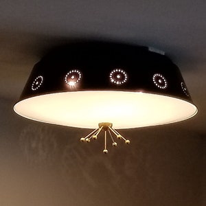 May include: A black ceiling light fixture with a white interior. The fixture has a circular shape with decorative dotted patterns around the edge. A cluster of gold-colored rods with spherical tips hangs from the center.