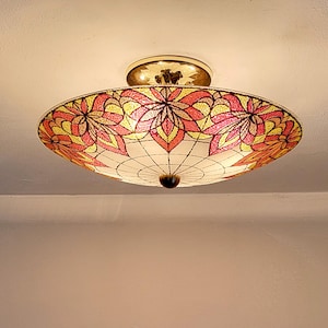May include: A circular flush mount ceiling light fixture featuring a stained glass shade with a floral pattern in shades of pink, yellow, and white. The fixture has a polished gold-tone metal base and a central dark brown finial.