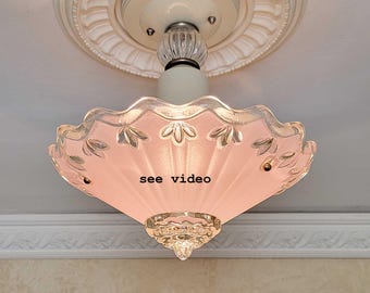 May include: A flush mount ceiling light fixture featuring a pale pink, scalloped glass shade with a floral embossed pattern. The fixture includes a white base, a clear glass finial at the bottom, and the text see video printed on the glass shade.
